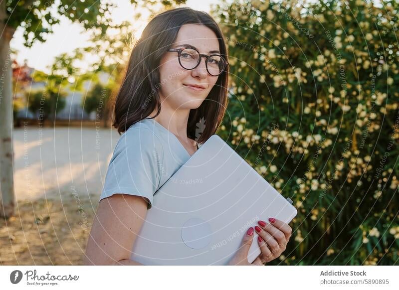 Junge Frau mit Laptop im Freien in sonniger Umgebung Baum Sonnenlicht Brille Lächeln Sommer Technik & Technologie Apparatur Gerät jung Erwachsener