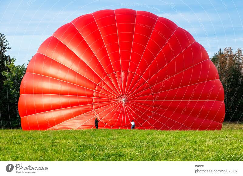 Heißluftballon in Rot, der vor dem Start auf einer Wiese am Waldrand mit heißer Luft gefüllt wird Ballonfahrer fliegen Bäume Luftverkehr vorher blau Windstille