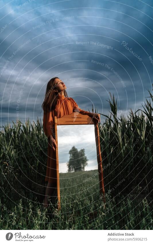 Frau in orangefarbenem Kleid mit Holzrahmen im Feld brünett Augen geschlossen orangefarbenes Kleid dramatischer Himmel Natur im Freien friedlich Windstille