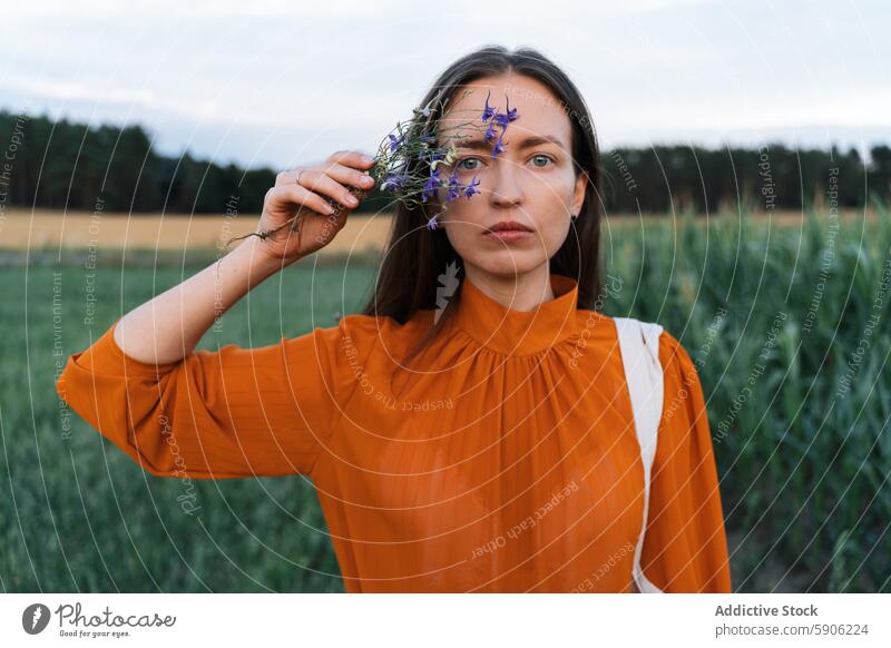 Brünette Frau hält Blumen auf einem Feld und schaut in die Kamera brünett Starrer Blick Fotokamera Gelassenheit orange Bluse grün Wildblume im Freien Natur