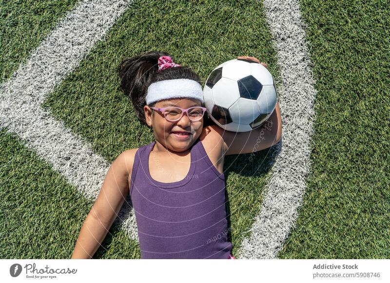 Lächelndes junges Mädchen hält einen Fußball auf dem Spielfeld Kind Ball Sport Feld Gras im Freien spielen Energie Glück Freude Brille Vielfalt Einschluss