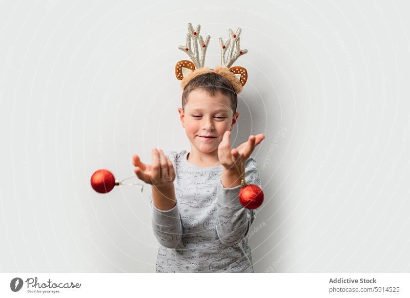 Ein junger Mann mit einem Rentier-Stirnband spielt mit Weihnachtsschmuck Weihnachten Junge Ornament festlich spielen werfen Freude Sprit Hintergrund rot