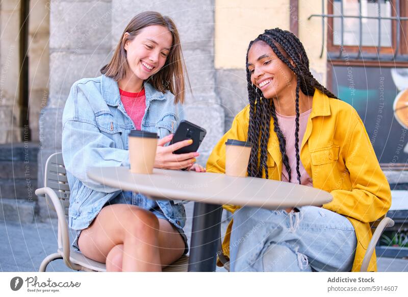 Zwei Frauen genießen Kaffee und Smartphone im Freien Freund Lächeln Café Tisch Sitzen Blick auf den Bildschirm rassenübergreifend vielfältig Gespräch