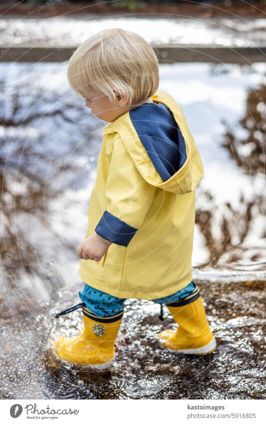 Kleiner blonder Junge in gelben Gummistiefeln und gelbem, wasserdichtem Regenmantel, der an einem bewölkten Regentag in Pfützen läuft. Kind im Regen. Säugling