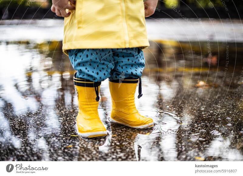 Kleiner Junge in gelben Gummistiefeln und gelbem, wasserdichtem Regenmantel, der an einem bewölkten Regentag in einer Pfütze steht. Kind im Regen. Säugling Spaß