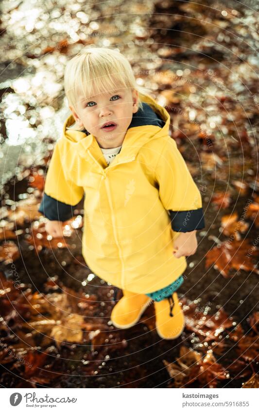 Kleiner blonder Junge in gelben Gummistiefeln und gelbem, wasserdichtem Regenmantel, der an einem bewölkten Regentag in Pfützen läuft. Kind im Regen. Säugling