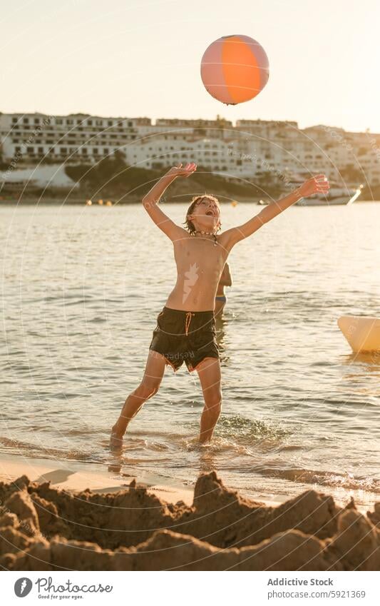 Glücklicher Junge spielt mit Ball am Ufer werfen MEER spielerisch Urlaub Sommer stehen Arme hochgezogen Wasser Strand Gebäude Ganzkörper spielen sorgenfrei