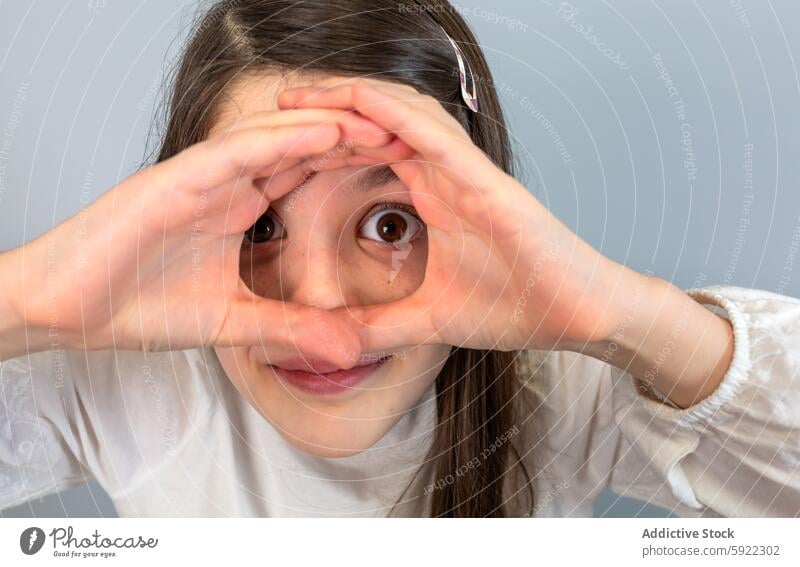 Ein junges Mädchen mit verspieltem Gesichtsausdruck schaut durch ein Fernglas, das sie mit ihren Händen vor einem grauen Hintergrund geformt hat. Kind