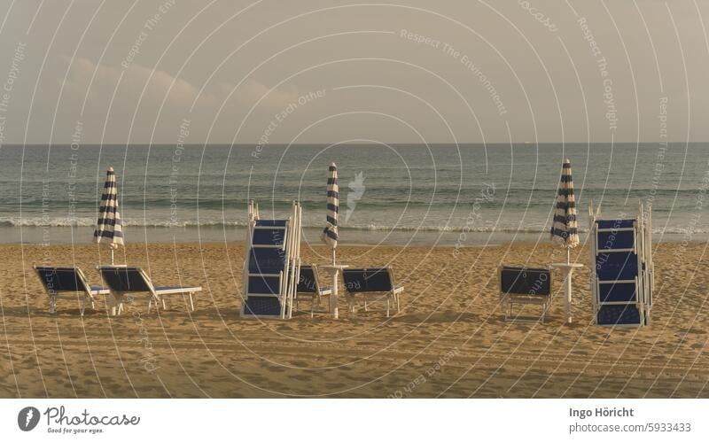 Unbenutzte blau-weiße Strandliege, zum Teil gestapelt, und 3 zusammengeklappte Sonnenschirme auf einem Sandstrand. Dahinter das Mittelmeer mit mäßigem Wellengang, ein verlassener Strand, menschenleer, das Foto ist sehr symmetrisch aufgebaut.