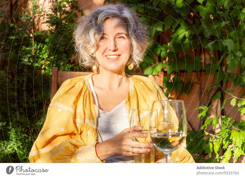 Reife Frau genießt Wein in einer Gartenanlage reif Glas im Freien Sonnenlicht Lächeln in die Kamera schauen freudig Sitzen Sommer Freizeit Erholung Laubwerk