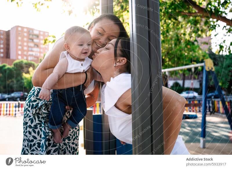 Mutter und Freundin küssen spielerisch ein Baby auf einem sonnigen Spielplatz Paar lesbisch lgbt kolumbianisch Küssen Freude im Freien Sonnenlicht Fröhlichkeit