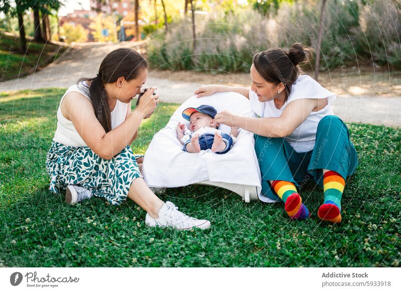 Zwei lesbische kolumbianische Frauen genießen einen sonnigen Tag im Park mit einem Baby auf einer Wiese lgbt Paar Gras im Freien Fotografie Familie Liebe Pflege