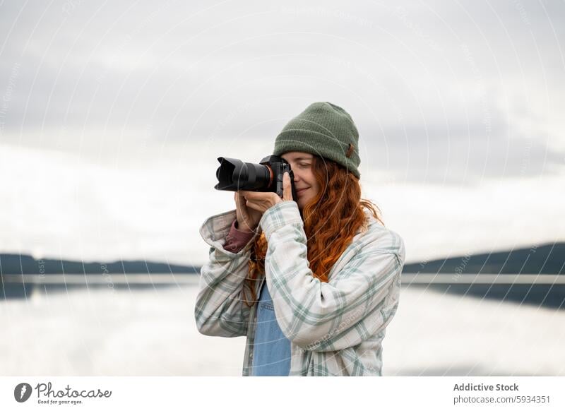 Frau, die während eines Campingausflugs Fotos am See macht Fotokamera Fotografie Landschaft im Freien ruhig rote Haare Beanie karierte Jacke Natur Hobby