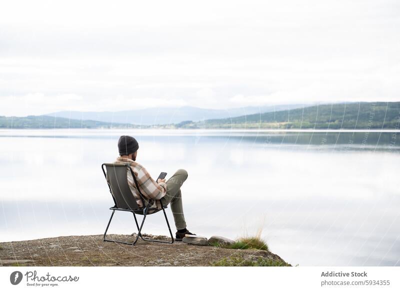 Mann genießt die Ruhe beim Camping am See mit einem Smartphone Stuhl Buch Gelassenheit ruhig Natur im Freien sitzend lesen Wald Berge u. Gebirge Nebel