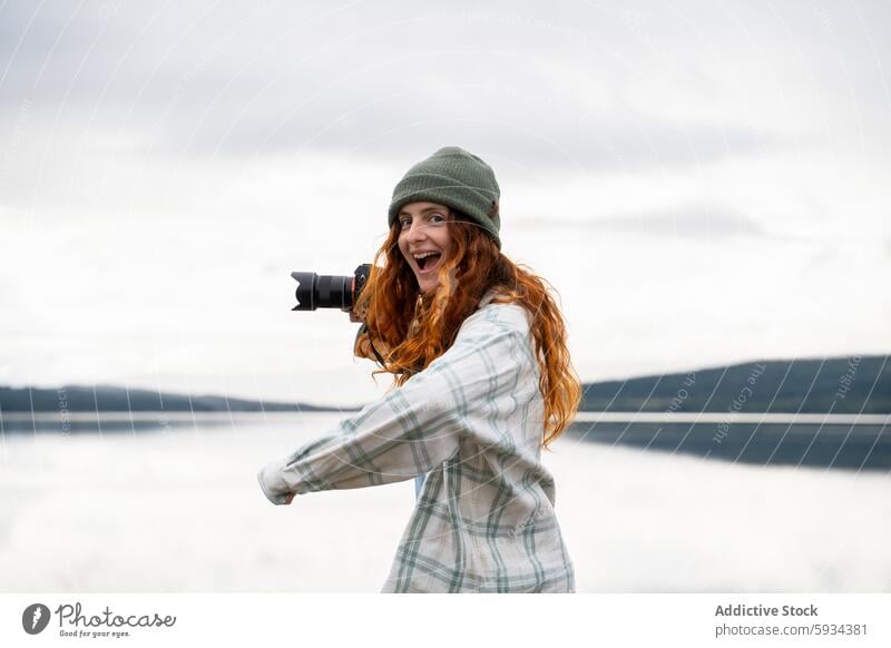 Glückliche Frau, die beim Camping am See Fotos macht Fotokamera Fotografie Natur Rotschopf heiter im Freien Fröhlichkeit malerisch Ansicht Beanie Plaid Hemd