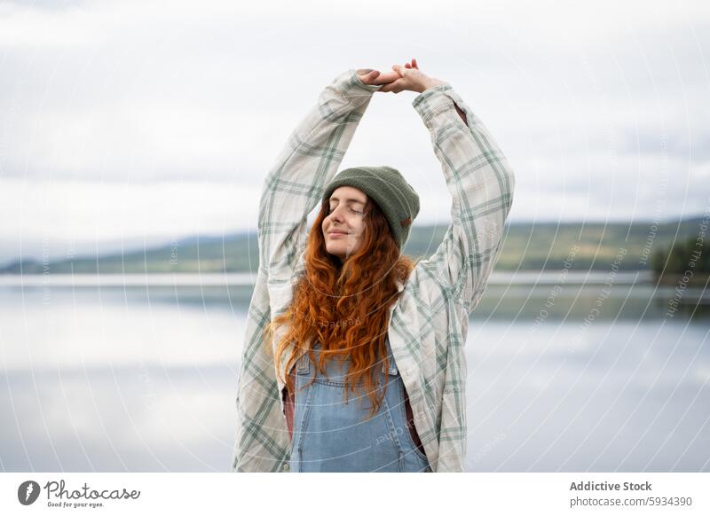 Frau genießt einen ruhigen Moment am See beim Camping Morgen Natur im Freien Dehnung Ruhe friedlich Flanellhemd Beanie Jeansstoff Latzhose Erholung Freude