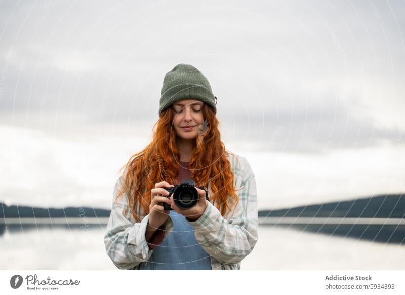 Junge Frau genießt die Fotografie am Seeufer Fotokamera Beanie rote Haare Camping Gelassenheit Natur friedlich im Freien kariertes Hemd Einsamkeit Windstille