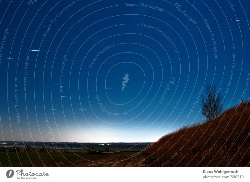 Weite Landschaft bei Nacht. Ein Weg führt in die Ferne und der Sternenhimmel wird von einem Kondensstreifen durchzogen. Lichter von Dörfern in der Ferne. Auf einem Ggrasbewachsenen Abhang steht ein kahler Baum.