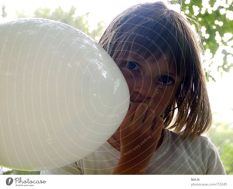 Nachdenkliches, verträumtes, süßes Mädchen mit weissem Luftballon in der Hand im Garten, schaut in die Kamera. Liebes Geburtstagskind draussen in der Natur freut sich über geschenkten Luftballon zu ihrem Fest, hält ihn fest und steckt ihn in den Mund.