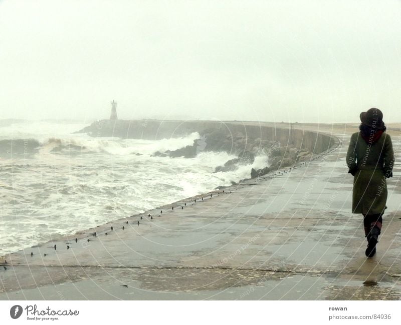 Spaziergang am Meer I überschwemmt See Frau Wellen Leuchtturm Gischt Nebel Pfütze Mantel grün gehen Bewegung unruhig Sturm nass kalt grau Wolken leer Wind