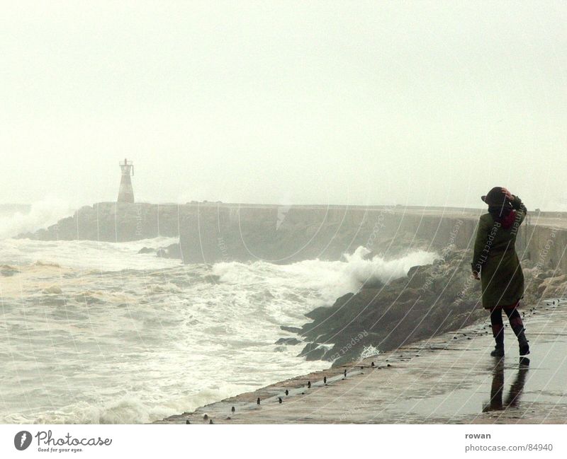 Spaziergang am Meer II überschwemmt See Frau Wellen Leuchtturm Gischt Nebel Pfütze Mantel grün gehen Bewegung unruhig Sturm nass kalt grau Wolken leer Wind