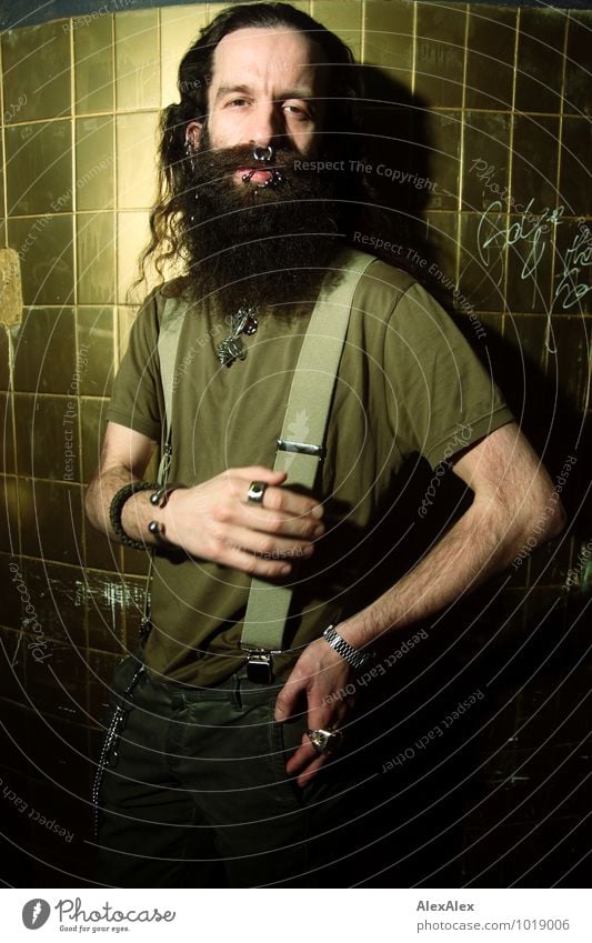 Young man with full beard and piercings stands in front of wall with golden tiles Room Man Adults 30 - 45 years T-shirt Suspenders Jewellery Piercing