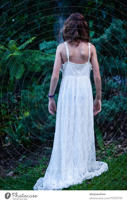 Girl in the garden with long white dress. She stands with her back to the camera, in front of her are green bushes and shrubs. Design Harmonious Garden Feminine