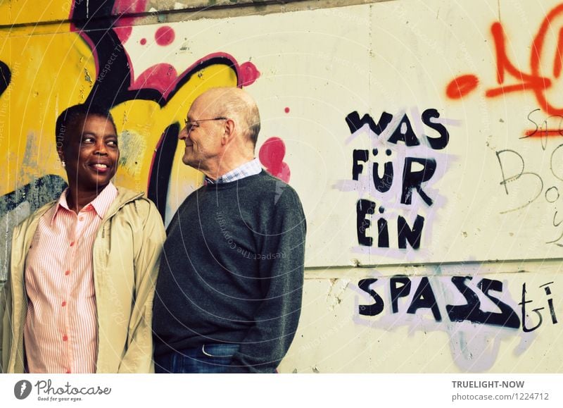 Beautiful coloured African woman and white senior form a couple with eye contact, standing close together in front of a wall painted with graffiti and the text WHAT FOR FUN, smiling lovingly into each other's eyes