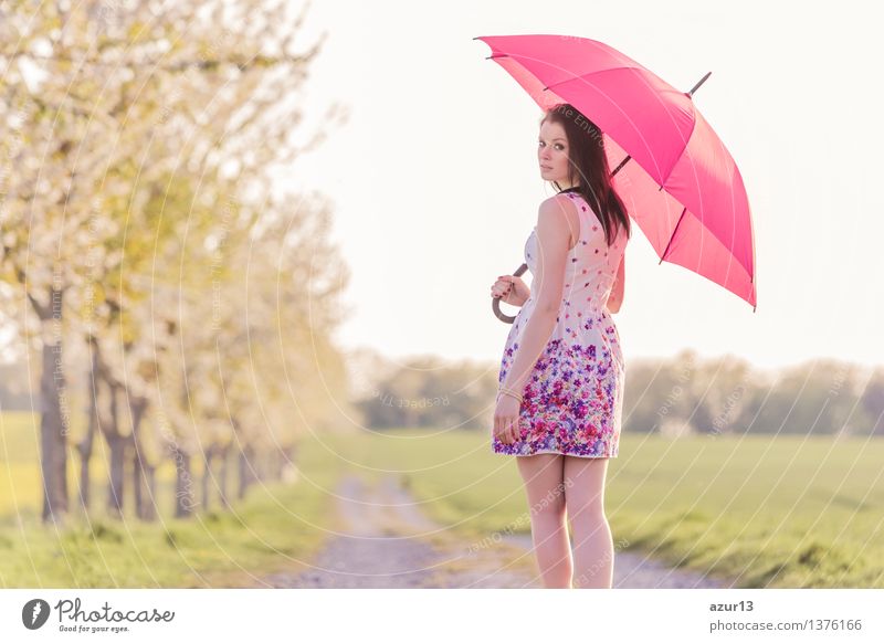 Woman with umbrella in changeable weather in spring or summer. The weather forecast is changeable between sun and rain. The attractive young woman goes for a walk on a path in the nature and takes a look back over her shoulder....