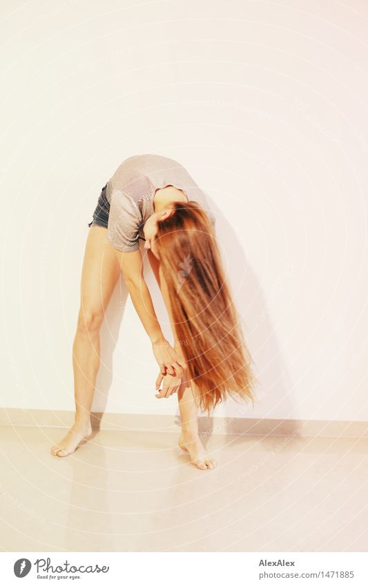 young, slim woman swings her upper body and her long, brunette hair in front of her legs, standing barefoot in front of a white wall Elegant Style pretty