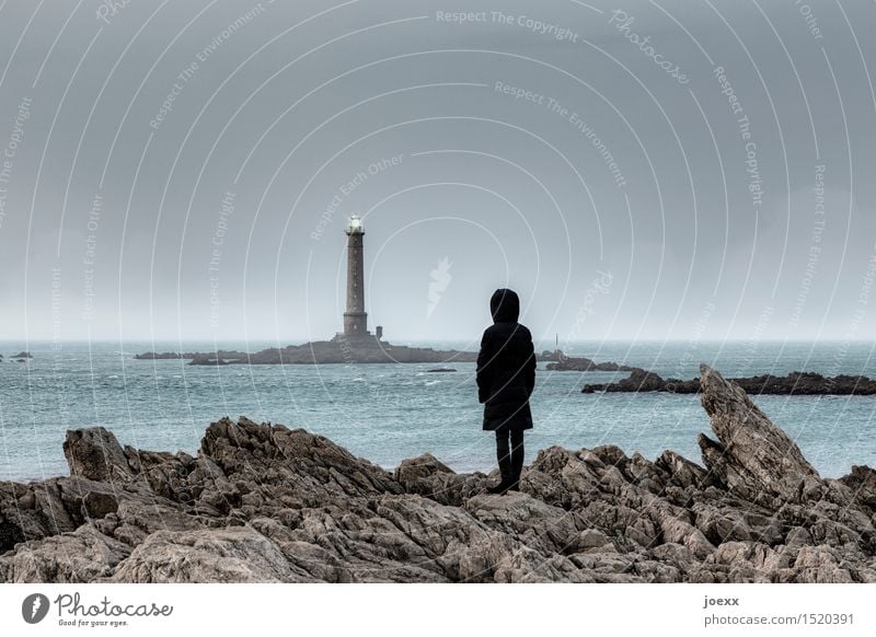 Woman in coat with hood observes distant lighthouse in sea Lighthouse Rock coast person rocky curt harsh somber Hope Longing Observe Ocean Water Horizon