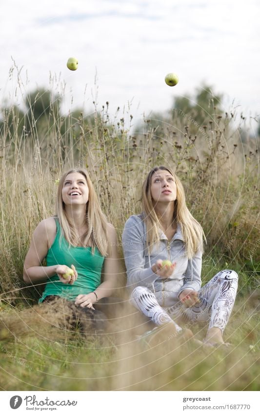 Two playful girls with long blond hair juggling outdoors as they sit side by side in long grass in the countryside Fruit Apple Happy Beautiful Wellness