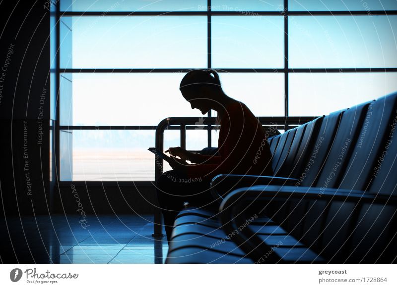 Woman in the aiport lounge sitting in chair by the window and using a tablet computer for checking her flight details with wifi connection. Silhouette Happy