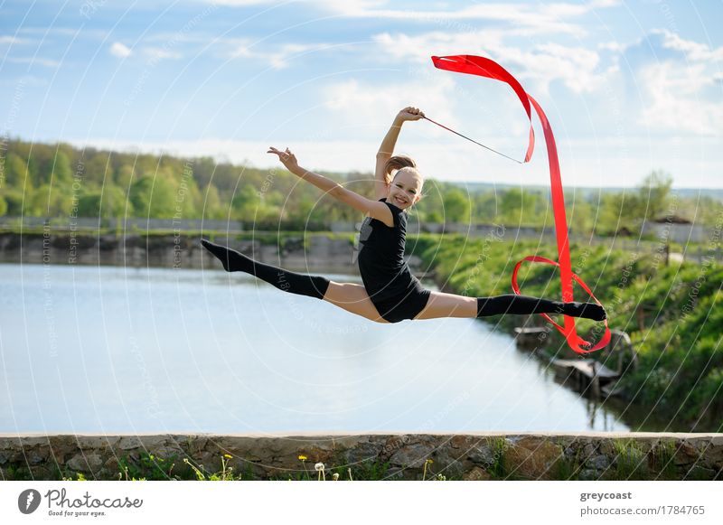 Rhythmic gymnast girl performing with a red ribbon outdoor. Leg-split in a jump against rural nature background Happy Body Summer Sports Success Child