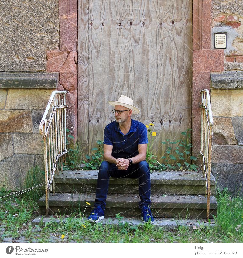 Man with beard, glasses and sun hat in dark blue clothes is sitting thoughtfully on a staircase in front of a dilapidated building Human being Masculine Adults