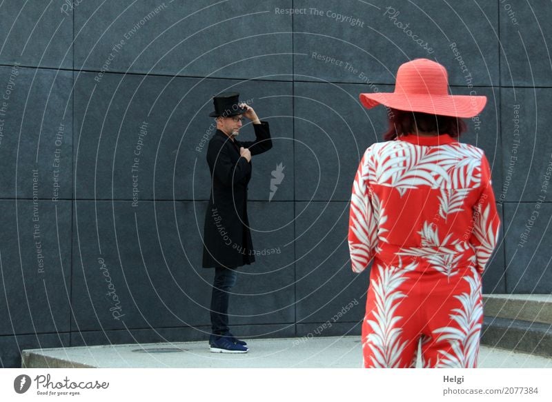 elegant gentleman in black frock coat and top hat is standing in front of a dark grey wall, in the foreground rear view of a woman with red-white overall and red hat