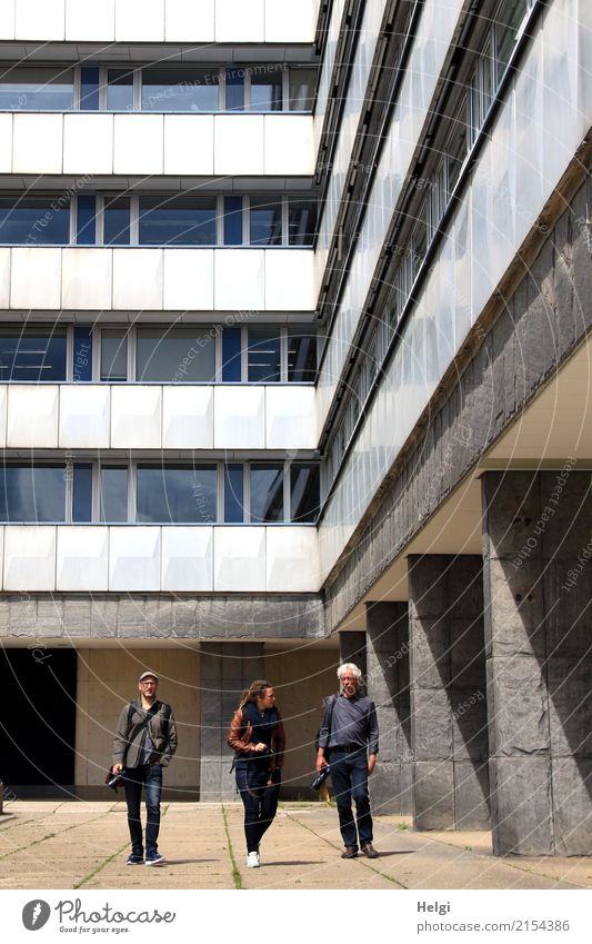 three persons, one woman and two men, walk on a footpath below a high-rise building Human being Masculine Feminine Woman Adults Male senior Man 3 45 - 60 years