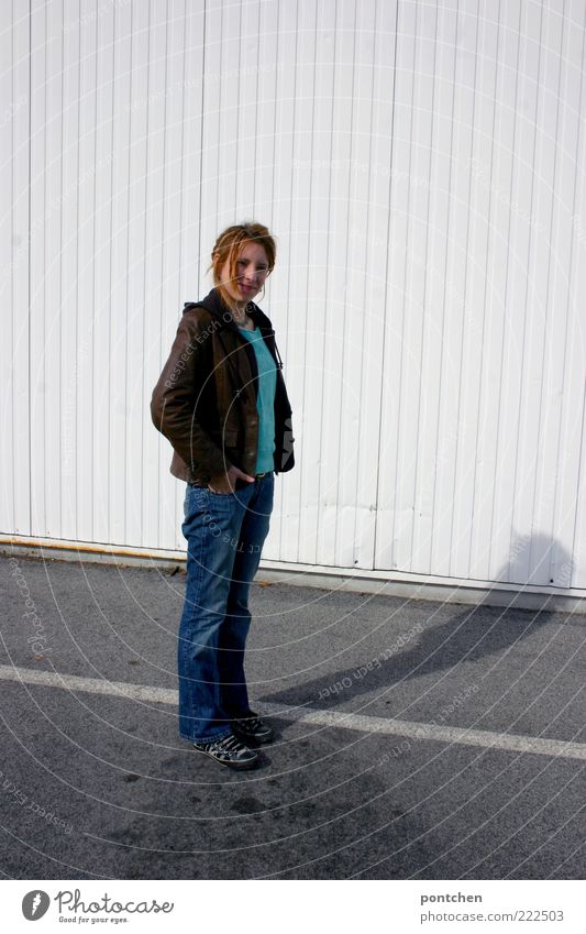 Young woman stands at the side of a white corrugated iron wall and casts shadows on the street and wall. Wait Youth (Young adults) Adults 1 Human being Jeans