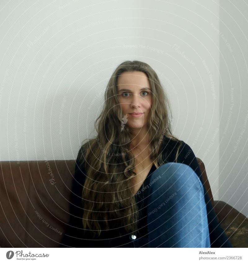 Portrait of young woman with long brunette hair and jeans pants sitting on brown sofa Style pretty Life Well-being Young woman Youth (Young adults) Face Knee