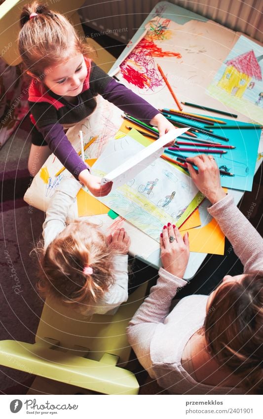 Little girl showing her picture her mom while she teaching children drawing a colorful pictures using pencil crayons sitting at table. Photo from above