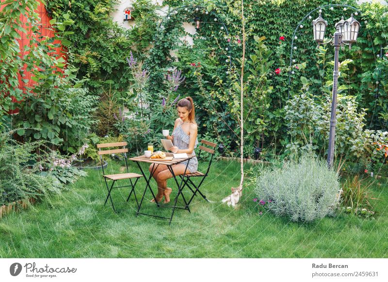 Young And Attractive Woman Having Morning Breakfast In Green Garden With French Croissant, Donuts, Coffee Cup, Orange Juice, Tablet and Notes Book On Wooden Table