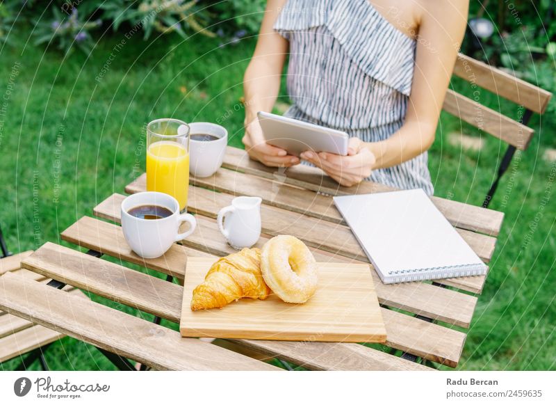 Young And Attractive Woman Having Morning Breakfast In Green Garden With French Croissant, Donuts, Coffee Cup, Orange Juice, Tablet and Notes Book On Wooden Table