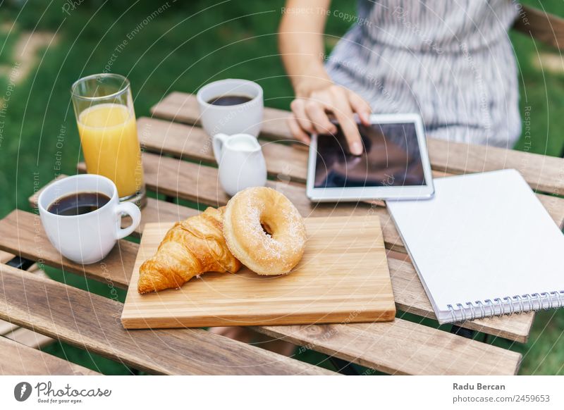 Young And Attractive Woman Having Morning Breakfast In Green Garden With French Croissant, Donuts, Coffee Cup, Orange Juice, Tablet and Notes Book On Wooden Table