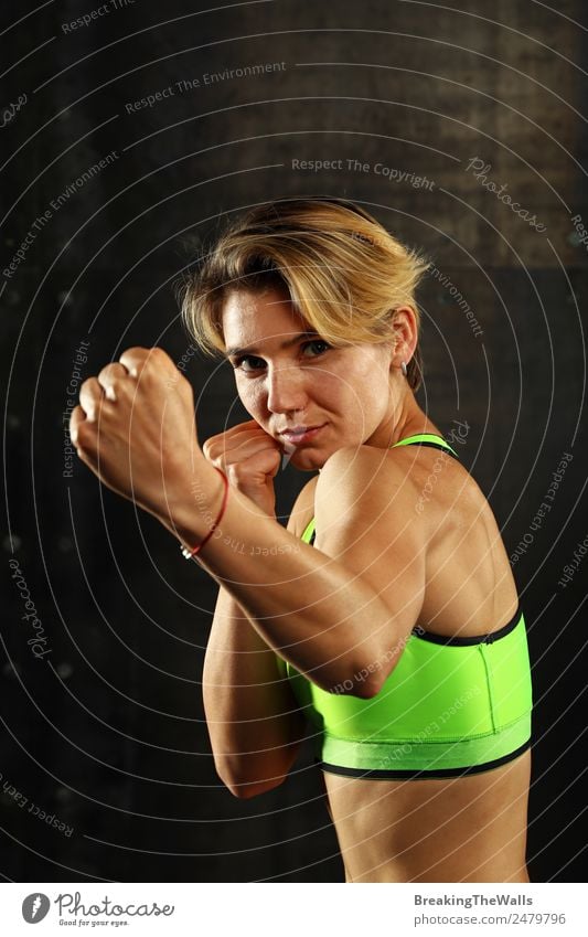 Close up front portrait of one young athletic woman in sportswear in gym over dark background, standing in boxing stance with hands and fists, looking at camera