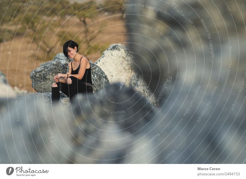 Beautiful young woman smiling sitting happy on the rocks outdoors in countryside attractive background beautiful black clothes brunette camera caucasian day