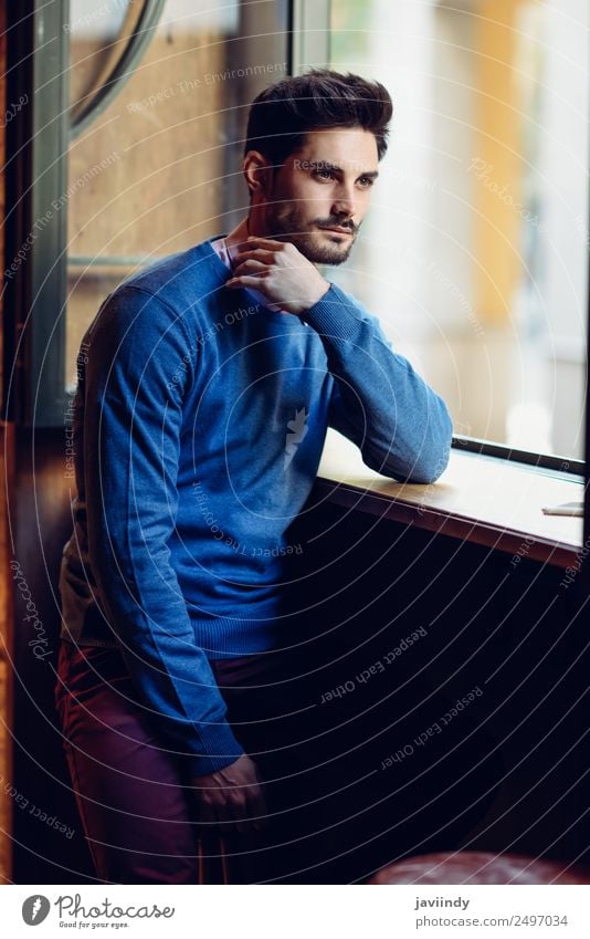 Thoughtful man with blue sweater with lost look near a window in a modern pub. Bearded guy with modern hairstyle Lifestyle Style Beautiful Hair and hairstyles