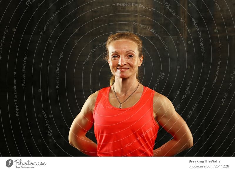 Close up front upper body portrait of one middle age athletic woman in sportswear in gym over dark background, looking at camera and smiling Lifestyle Sports