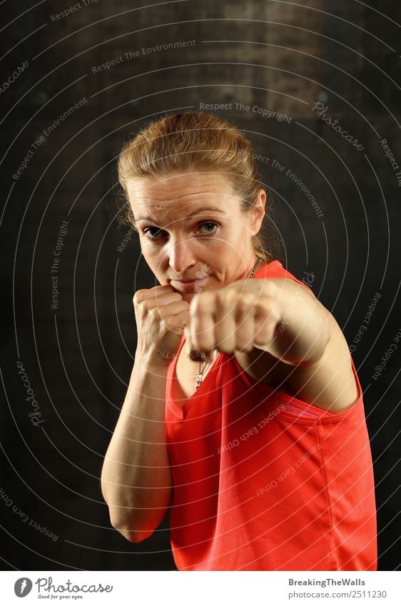 Close up front portrait of one young mid adult athletic woman in sportswear in gym over dark background, standing in boxing stance with hands and fists, looking at camera