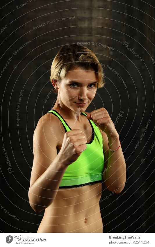 Close up front portrait of one young athletic woman in sportswear in gym over dark background, standing in boxing stance with hands and fists, looking at camera