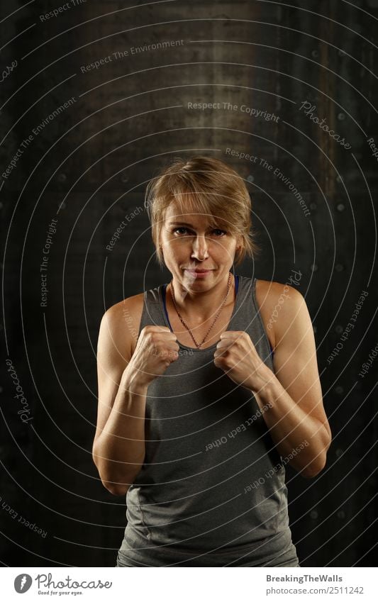 Close up front portrait of one young middle age athletic woman in sportswear in gym over dark background, standing in boxing stance with hands and fists, looking at camera